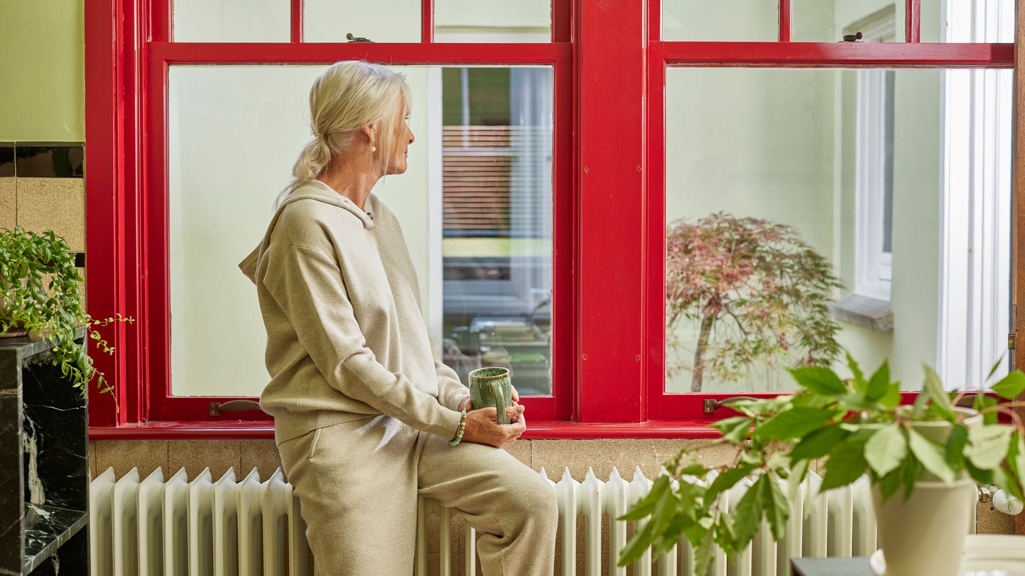 Donna seduta su un termosifone di fianco a una grande finestra, che si gode il calore di una casa a basso consumo energetico