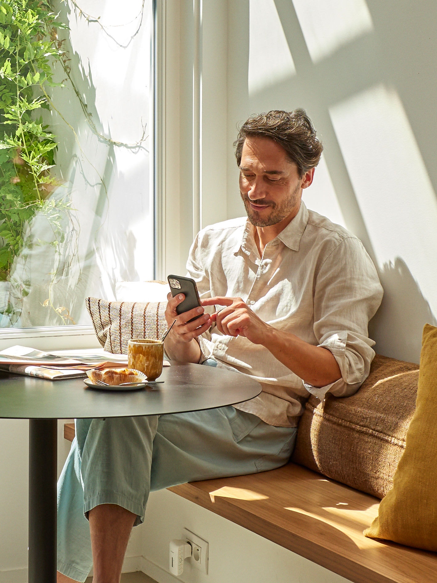 Un uomo si rilassa e guarda il telefono mentre è seduto a tavola davanti a una tazza di caffè e qualcosa da mangiare