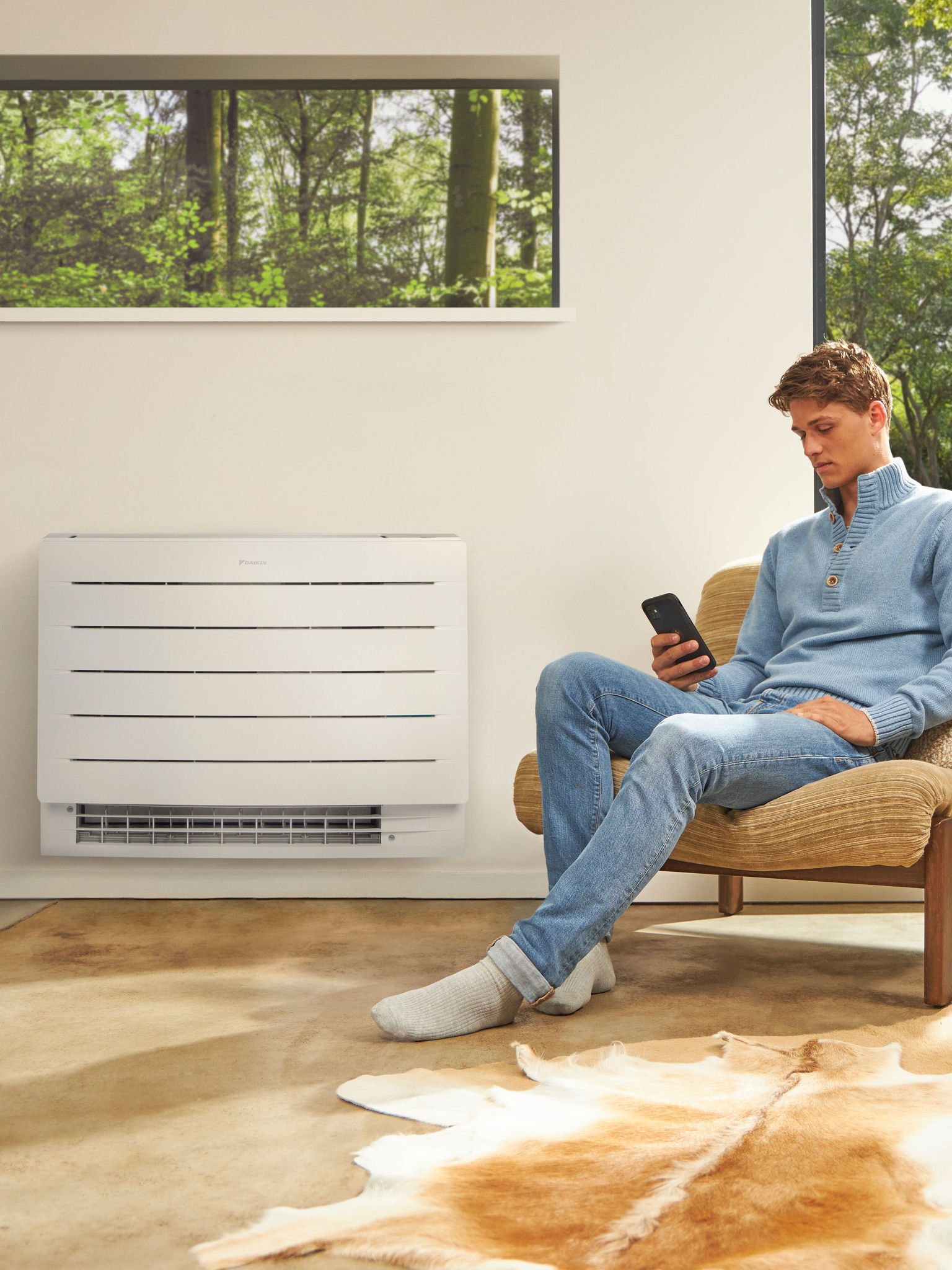 Un uomo seduto in una stanza luminosa con una pompa di calore installata sulla parete alle sue spalle guarda il suo smartphone
