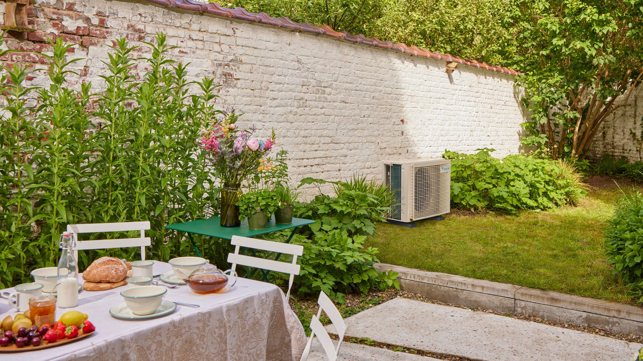 Tavola apparecchiata per una colazione in giardino con vista su un climatizzatore a Inverter che raffresca una casa adiacente