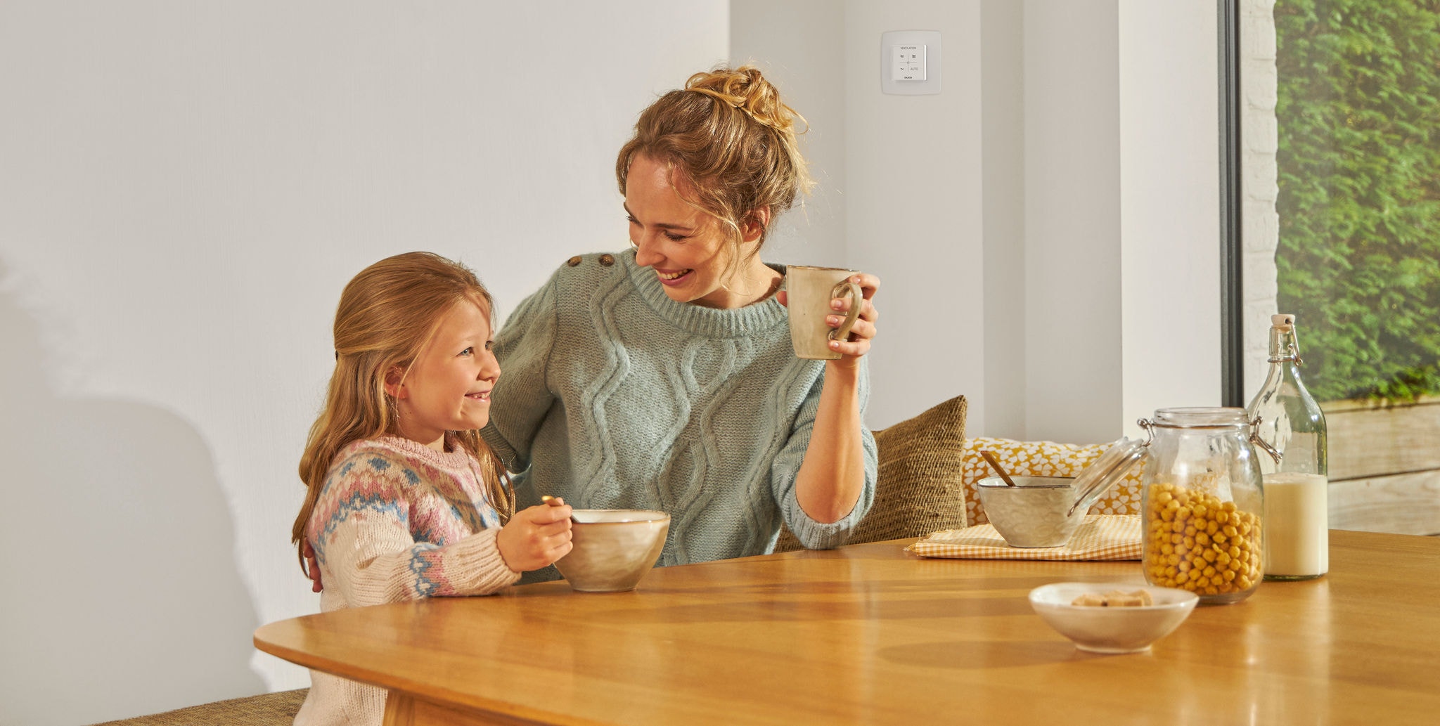 Una ragazzina mangia seduta a tavola accanto a una donna con una tazza in mano; entrambe sorridono, serene e rilassate, e si godono un’aria pulita.