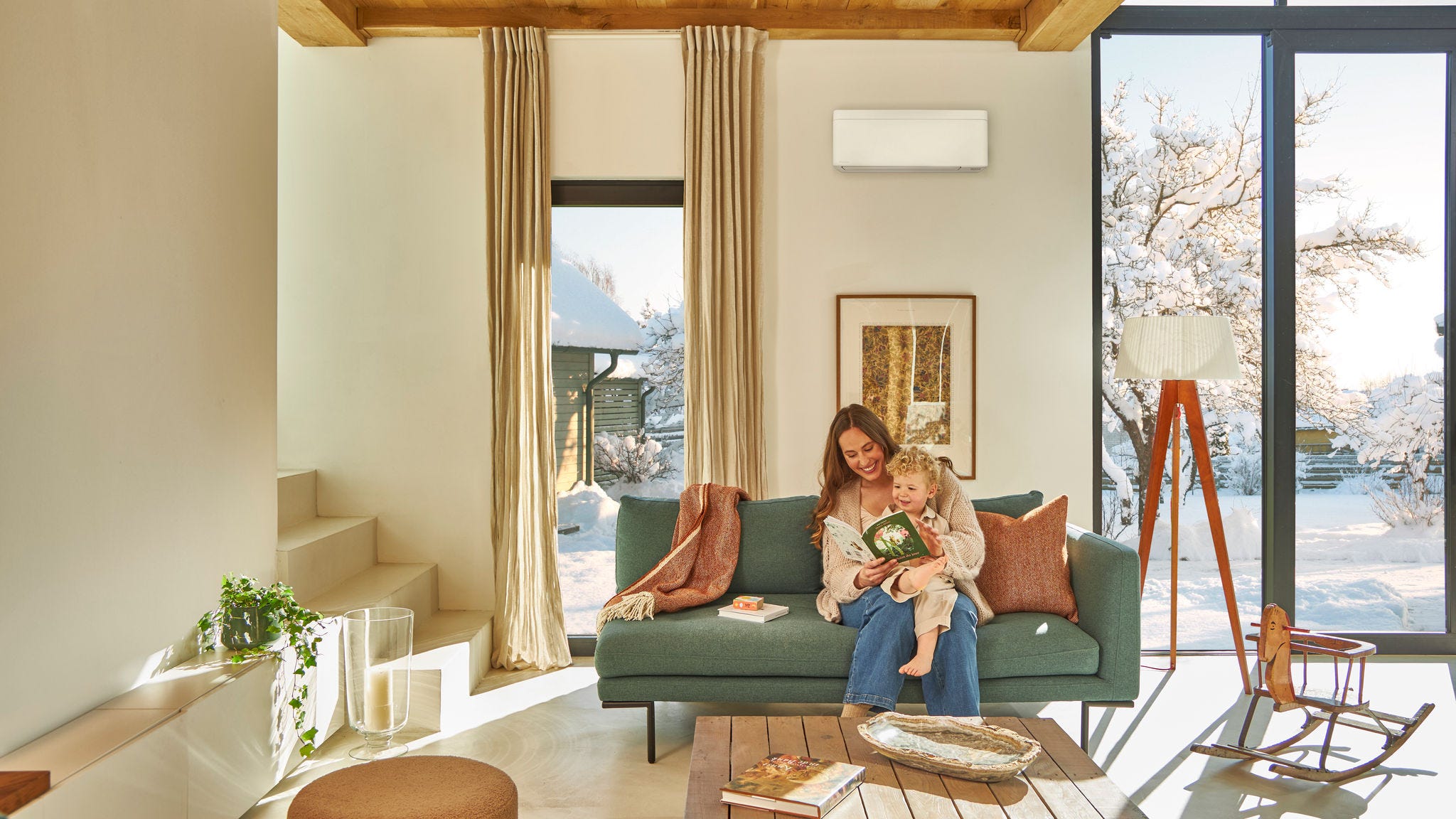 Una mamma seduta sul divano sotto una pompa di calore aria-aria a parete legge un libro per bambini con il suo figlioletto in grembo godendosi il caldo mentre fuori nevica